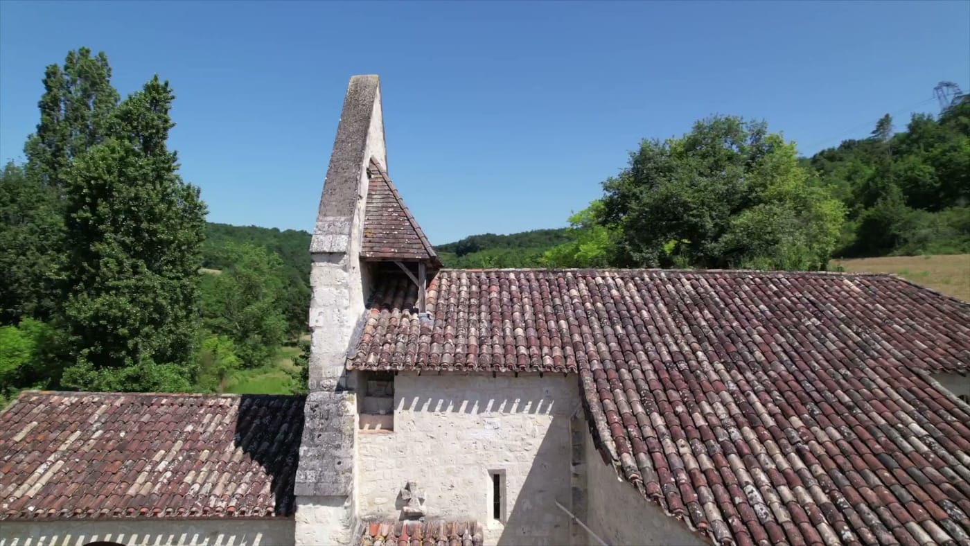 L'église de Cabalsaut vue du ciel : église romane en pierre claire coiffée d'un clocher-mur triangulaire et d'une toiture de tuiles anciennes, une croix de pierre sur le mur, entourée d'arbres dans le vallon.