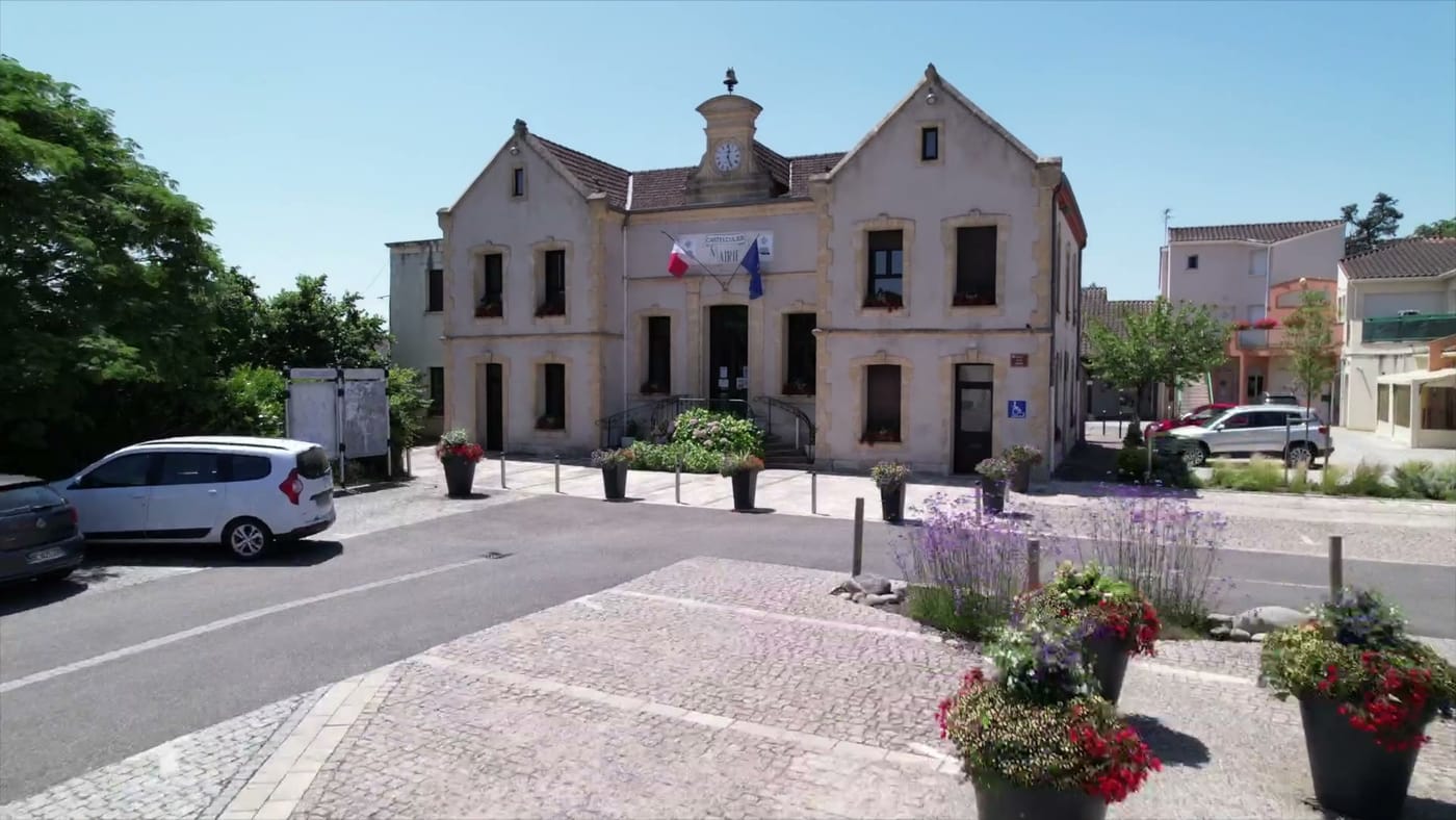 La mairie de Castelculier vue depuis la place : bâtiment en pierre claire à deux pignons, horloge et clocheton au centre, drapeaux français et européen encadrant l'entrée, massifs de fleurs sur la place pavée.