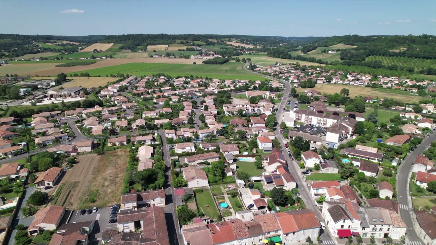 Vue aérienne du village de Castelculier : maisons aux toits de tuiles regroupées au creux d'une vallée, entourées de champs cultivés et de coteaux boisés à l'horizon.