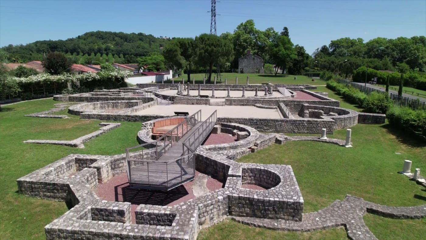 Vue aérienne du site archéologique de Villascopia : fondations en pierre et alignements de colonnes d'une villa gallo-romaine, parcourus par une passerelle de visite métallique, au milieu d'une vaste pelouse.