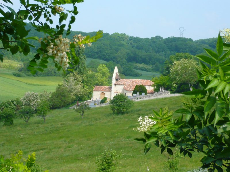 L'église de Cabalsaut vue depuis les prés du vallon : le clocher-mur et la nef aux toits de tuiles entre les arbres, le petit cimetière au premier plan, les coteaux boisés derrière.