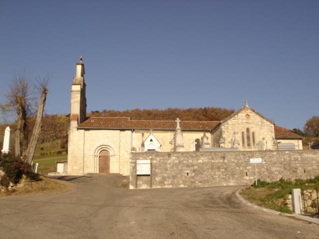 L'église de Saint-Amans en pierre blonde sous un ciel bleu d'automne : clocher-tour à arcades sur la gauche, portail roman en plein cintre, pignon percé de baies, cimetière ceint d'un mur au premier plan.