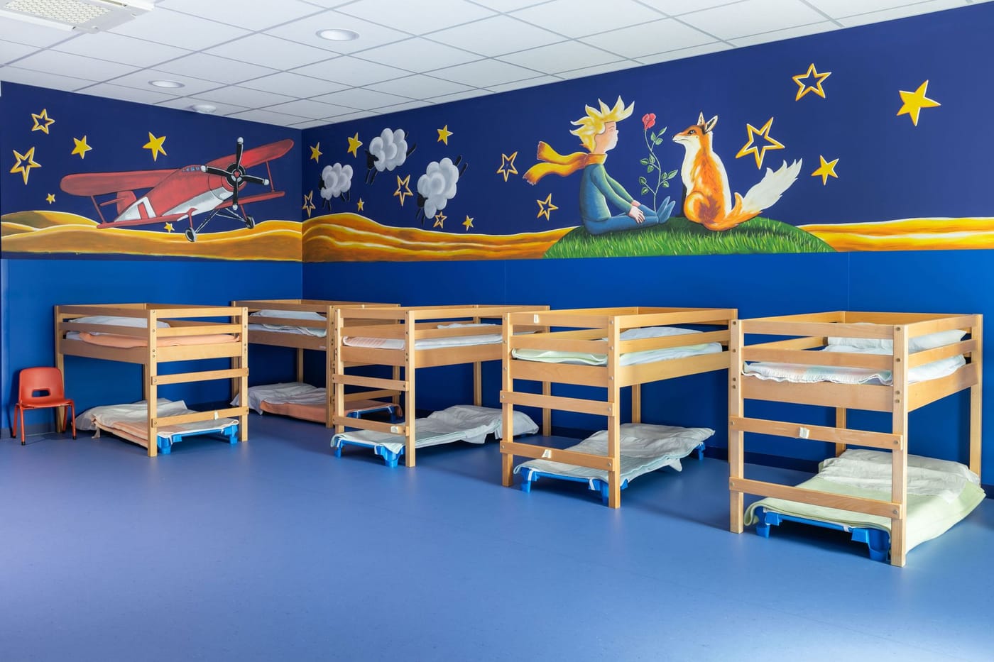 Dortoir aux murs bleus décoré d'une fresque du Petit Prince ; lits superposés en bois alignés pour la sieste.