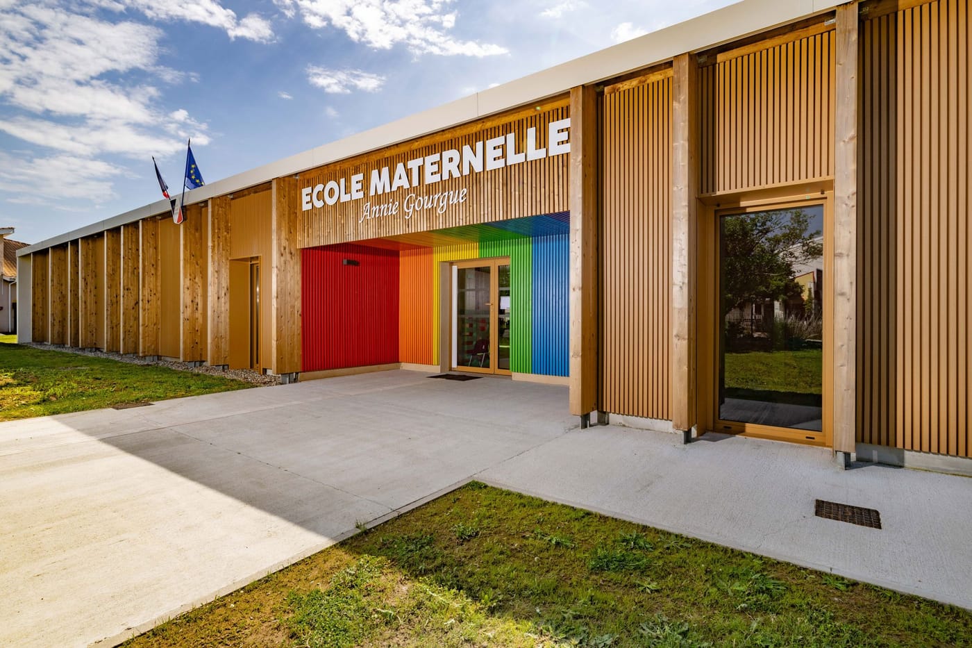 Façade de l'école maternelle Annie Gourgue : long bâtiment bas en bardage de bois clair, dont l'entrée centrale est habillée de lames aux couleurs de l'arc-en-ciel.