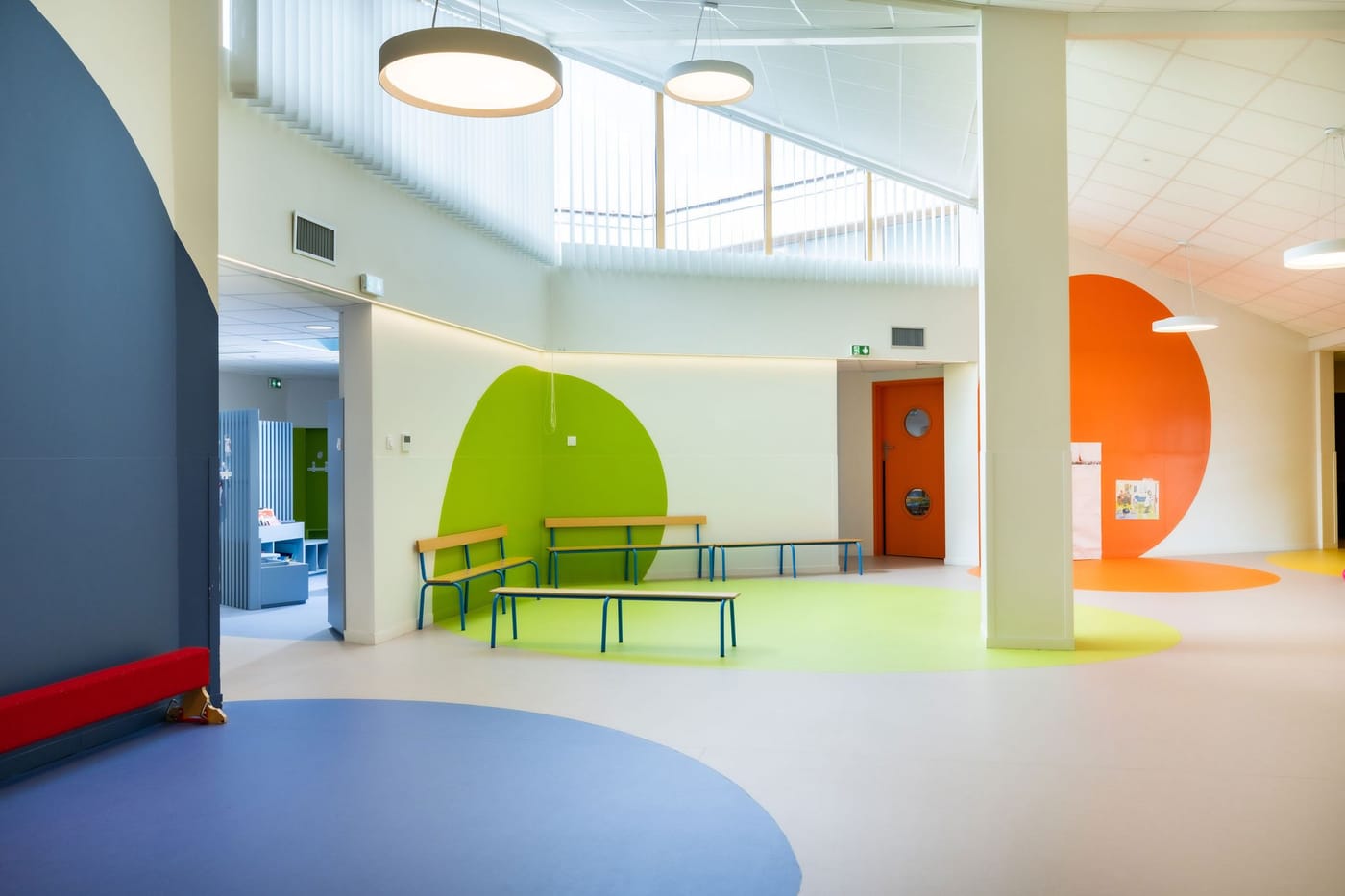 Hall intérieur lumineux et coloré : grands cercles peints en vert et orange au sol et au mur, bancs bas pour les enfants.
