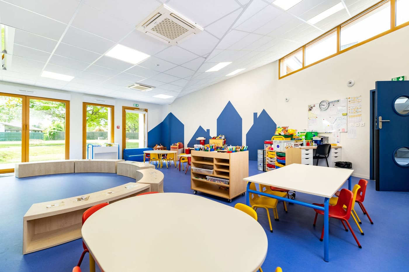 Salle de classe spacieuse avec tables rondes, chaises colorées et meubles de rangement, ouverte sur l'extérieur par de larges baies vitrées.