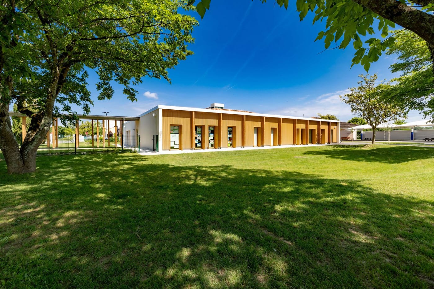 L'école maternelle vue depuis la pelouse arborée qui l'entoure, sous un ciel bleu.