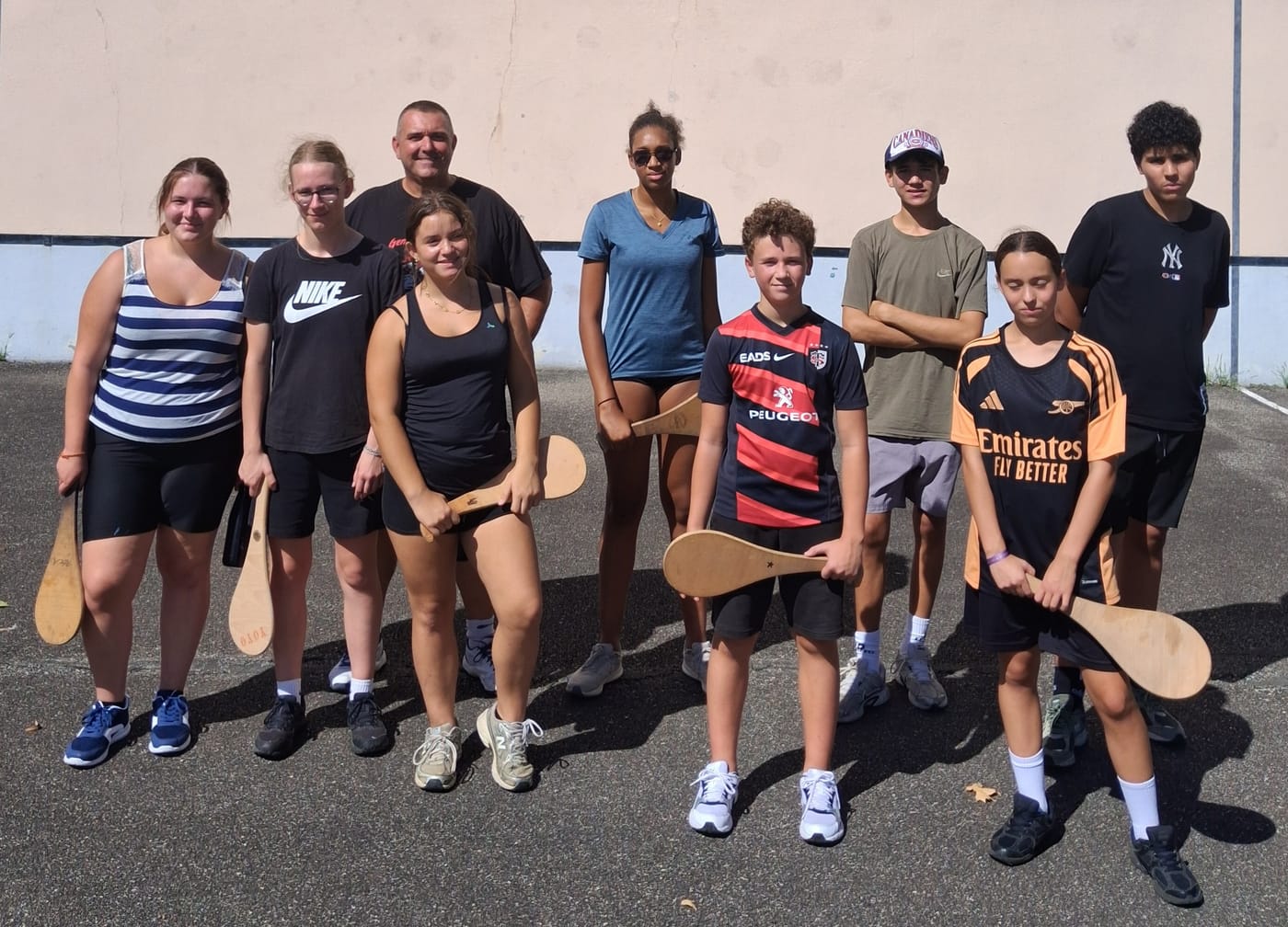 Un groupe du chantier citoyen aligné sur un terrain de sport avec leur animateur, chacun une raquette de bois à la main, devant le mur d'un fronton.