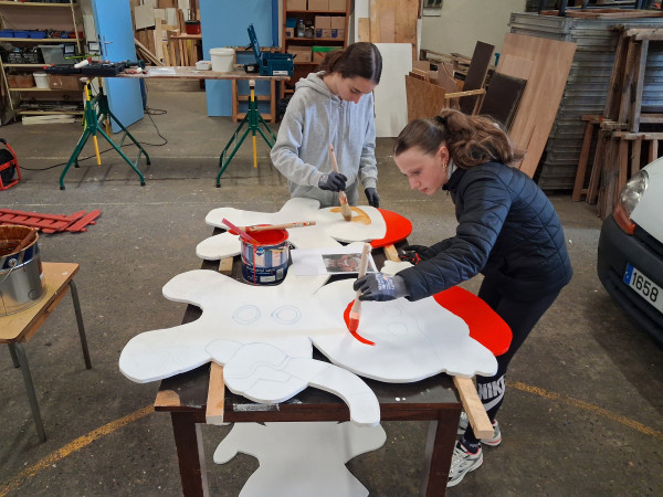 Deux adolescentes peignent en blanc et rouge de grandes silhouettes en bois découpé, à l'atelier municipal.