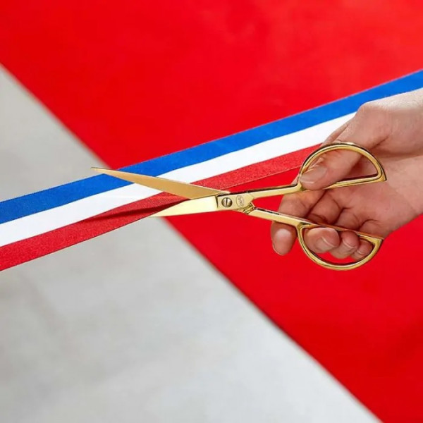 Une main coupe un ruban tricolore à l'aide de ciseaux dorés, sur un tapis rouge — symbole d'inauguration.