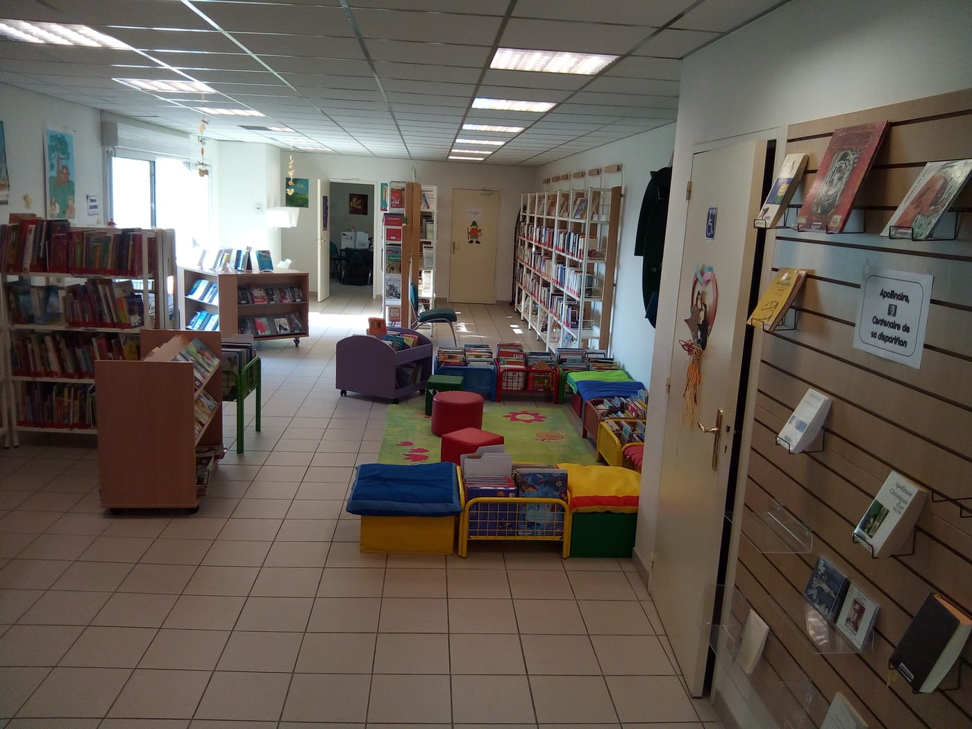 L'intérieur de la bibliothèque : rayonnages de livres le long des murs, tapis fleuri et petits fauteuils colorés du coin jeunesse au centre, présentoirs de nouveautés à droite.